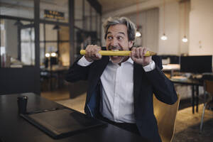 Wütender älterer Geschäftsmann mit riesigem Bleistift am Schreibtisch im Büro - GUSF02774