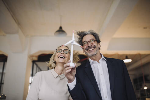 Happy businessman and businesswoman holding wind turbine model in office - GUSF02781