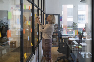 Mature businesswoman writing on adhesive notes on glass pane in office - GUSF02793