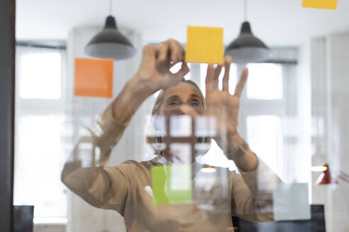 Mature businesswoman with adhesive notes on glass pane in office - GUSF02794