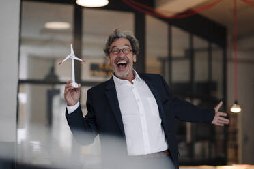 Excited senior businessman with wind turbine model in office - GUSF02804