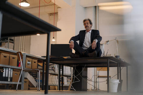 Senior businessman sitting cross-legged on desk in office meditating - GUSF02811