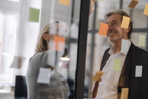 Glücklicher Geschäftsmann und Geschäftsfrau arbeiten an Haftnotizen an einer Glasscheibe im Büro, lizenzfreies Stockfoto