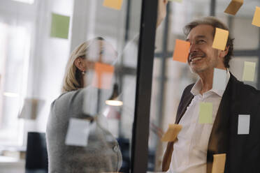 Happy businessman and businesswoman working on sticky notes at glass pane in office - GUSF02832