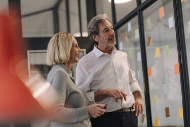 Businessman and businesswoman working on sticky notes at glass pane in office - GUSF02842