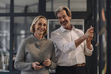 Happy businessman and businesswoman working on sticky notes at glass pane in office - GUSF02843