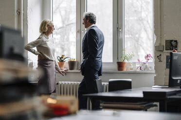 Smiling businessman and businesswoman talking at the window in office - GUSF02909