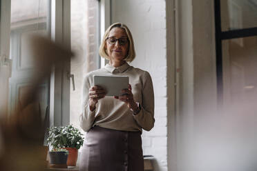 Mature businesswoman using tablet at the window in office - GUSF02920