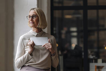 Mature businesswoman holding tablet at the window in office - GUSF02921