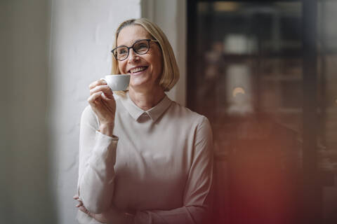 Lächelnde reife Geschäftsfrau, die eine Kaffeepause am Fenster im Büro macht, lizenzfreies Stockfoto