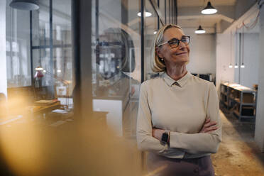 Portrait of smiling mature businesswoman leaning against glass pane in office - GUSF02936