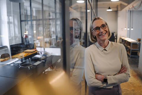 Porträt einer lächelnden reifen Geschäftsfrau, die sich gegen eine Glasscheibe im Büro lehnt, lizenzfreies Stockfoto