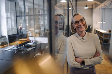 Porträt einer lächelnden reifen Geschäftsfrau, die sich gegen eine Glasscheibe im Büro lehnt - GUSF02937