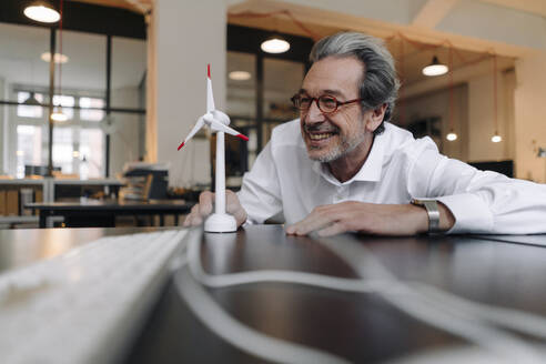Happy senior businessman with wind turbine model in office - GUSF02938