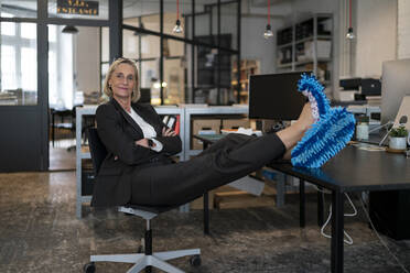 Reife Geschäftsfrau mit Füßen auf dem Schreibtisch in Reinigungspantoffeln im Büro - GUSF02939