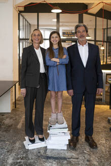 Portrait of smiling businessman and businesswoman with girl standing on stack of books in office - GUSF02942