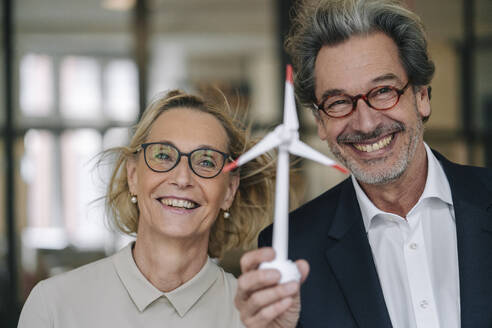 Portrait of happy businessman and businesswoman holding wind turbine model in office - GUSF02950