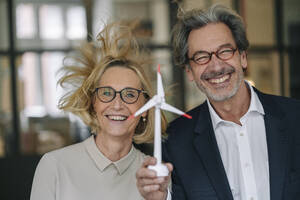 Portrait of happy businessman and businesswoman holding wind turbine model in office - GUSF02951