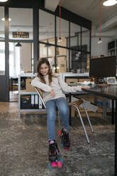 Girl with roller skates sitting at desk in office wobbling with chair - GUSF02964