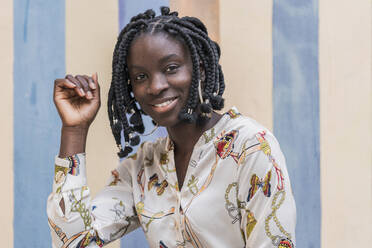 Portrait of smiling young woman with black braids in front of striped wall - DLTSF00295