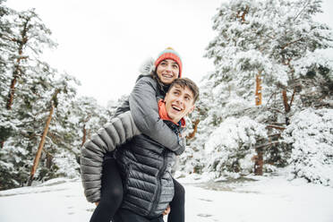 Portrait of happy young man giving his girlfriend a piggyback ride in winter forest - OCMF00927