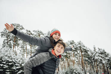 Porträt eines lächelnden jungen Mannes, der seine glückliche Freundin im Winterwald huckepack nimmt - OCMF00928