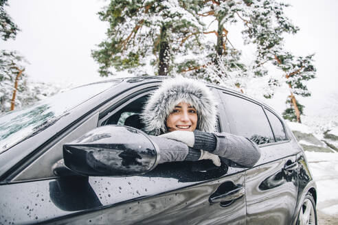 Portrait of smiling young woman leaning out of car window in winter forest - OCMF00933