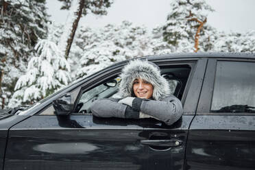 Porträt einer lächelnden jungen Frau, die sich im Winterwald aus dem Autofenster lehnt - OCMF00936