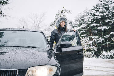 Smiling young woman standing beside parked car in winter forest looking at distance - OCMF00937