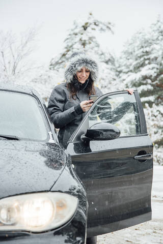 Smiling young woman standing beside parked car in winter forest looking at cell phone stock photo