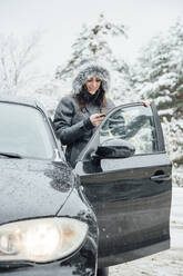 Smiling young woman standing beside parked car in winter forest looking at cell phone - OCMF00938