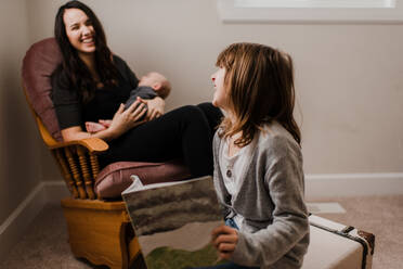 Mädchen lacht mit Mutter, während sie ihren kleinen Bruder im Wohnzimmersessel wiegt - ISF22991