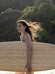 Junge Surferin mit langen, vom Wind zerzausten Haaren, die ein Surfbrett am Strand trägt, Victoria, Australien - ISF23024