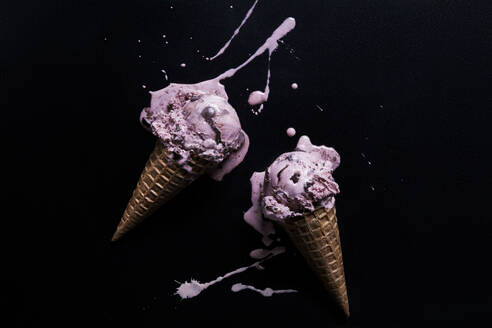 Two chocolate chip raspberry ice-cream cones with melted splash against black background, overhead view - ISF23056