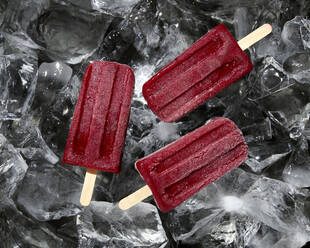 Three homemade raspberry iced lollies melting on chunks of ice, overhead view - ISF23068