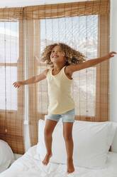 Little girl jumping with arms open on bed in beach house - ISF23134
