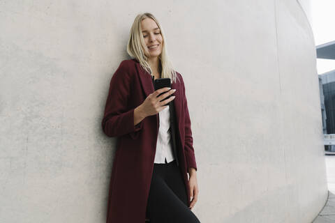 Blonde Geschäftsfrau, die ein Smartphone benutzt und sich an eine Wand lehnt, lizenzfreies Stockfoto