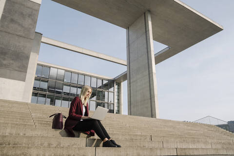 Blonde Geschäftsfrau mit Laptop im Hintergrund eines modernen Bürogebäudes, lizenzfreies Stockfoto