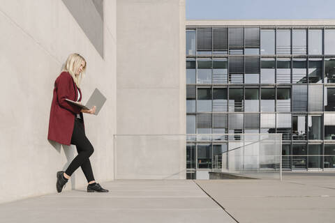 Blonde Geschäftsfrau, die einen Laptop benutzt und sich an eine Wand lehnt, lizenzfreies Stockfoto