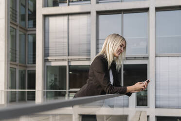 Blonde Geschäftsfrau mit Smartphone im Hintergrund modernen Gebäude - AHSF01383