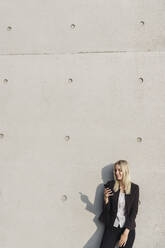 Blonde Geschäftsfrau mit Smartphone, die sich an eine Wand lehnt und nach unten schaut - AHSF01388