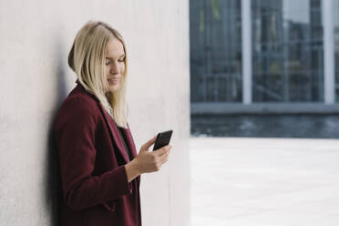 Blond businesswoman using smartphone, leaning on a wall, looking down - AHSF01395