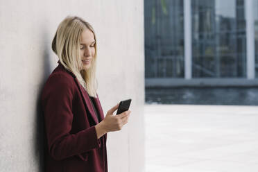 Blonde Geschäftsfrau, die ein Smartphone benutzt, an eine Wand gelehnt, mit Blick nach unten - AHSF01395