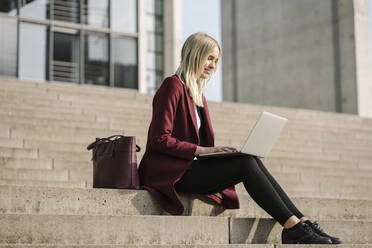 Blond businesswoman using laptop, sitting on step - AHSF01399