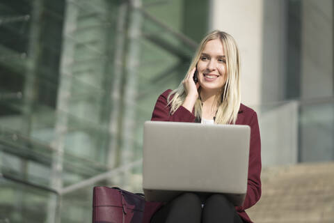 Blonde Geschäftsfrau, die einen Laptop benutzt und telefoniert, sitzt auf einer Treppe, lizenzfreies Stockfoto