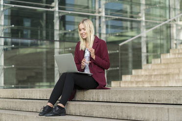 Blond businesswoman using laptop and drinking from water bottle, sitting on steps - AHSF01403