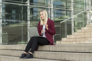 Blonde Geschäftsfrau, die einen Laptop benutzt und aus einer Wasserflasche trinkt, sitzt auf einer Treppe - AHSF01403