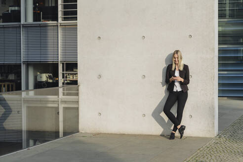 Blonde Geschäftsfrau mit Smartphone an die Wand gelehnt - AHSF01408