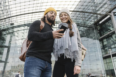 Glückliches junges Paar mit einem Smartphone am Hauptbahnhof, Berlin, Deutschland - AHSF01416