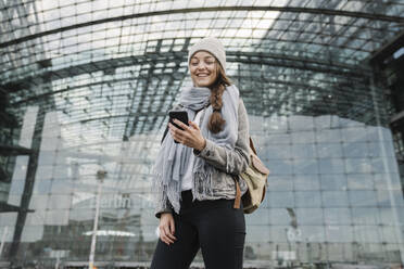Glückliche junge Frau mit Smartphone am Hauptbahnhof, Berlin, Deutschland - AHSF01420